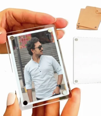 A close-up view of a hand holding a vertical rectangle-shaped personalized acrylic magnetic fridge magnet frame containing a photo of a man in sunglasses, with a separate blank clear block and a brown paper covered piece in the background by upahar18.