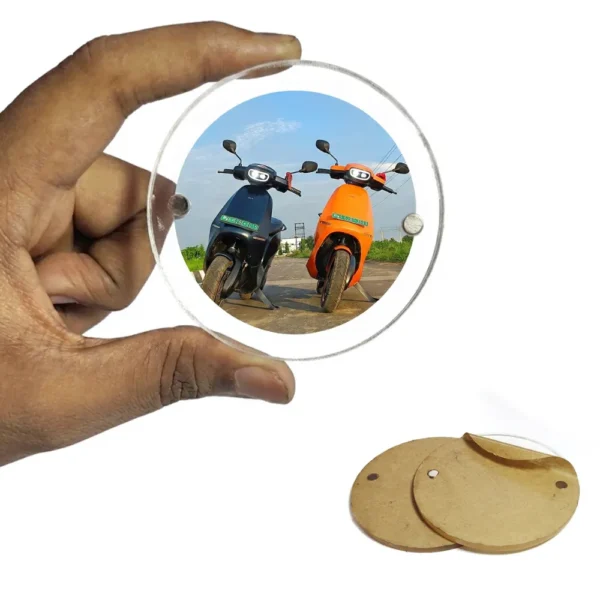 A hand holding a circular round-shaped clear acrylic magnetic frame featuring a personalized photo of two modern electric scooters on a sunny road, next to two unpeeled brown paper heart sheets by upahar18.