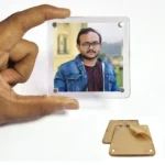 A person holding a square-shaped borderless acrylic magnetic photo frame showing a customized portrait of a man in a denim jacket and glasses, with a pair of protective brown kraft paper layers beside it by upahar18.