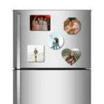 A straight front view of a stainless steel refrigerator door holding a complete combo set of five different shaped custom MDF photo fridge magnets, including a rectangle frame with four women in ethnic dresses, an octagon frame with an elderly couple, a round frame with two women posing, a square frame with a man walking on a beach, and a heart-shaped frame showing a woman with puppies, all featuring vibrant personalized photos by upahar18.