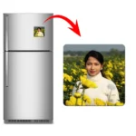 A product presentation featuring a double-door steel refrigerator on the left with a square personalized flexible fridge magnet attached to it. A large red curved arrow points from the fridge to a close-up view of the square magnet on the right, displaying a customized photo of a smiling young woman in a white turtleneck sweater standing in a vibrant field of yellow mustard flowers by upahar18.