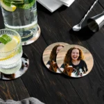 A flat-lay view on a dark rustic wooden table featuring refreshing lime mocktail glasses, bar tools, and circular MDF coasters from upahar18 printed with a personalized custom photo of a smiling woman and her pet dogs.