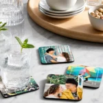 A luxury lifestyle setup on a white marble table showing iced water glasses, a bowl of pistachios, and four square MDF coasters from upahar18 printed with personalized custom photos of couples and geometric patterns.