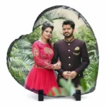 A heart shape natural rock stone frame from upahar18, displaying a personalized custom photo printed on its glossy surface. The printed image features a smiling couple in traditional South Asian wedding attire standing against a lush green garden background, supported by two black stands over a plain white background.
