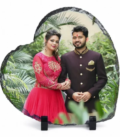 A heart shape natural rock stone frame from upahar18, displaying a personalized custom photo printed on its glossy surface. The printed image features a smiling couple in traditional South Asian wedding attire standing against a lush green garden background, supported by two black stands over a plain white background.