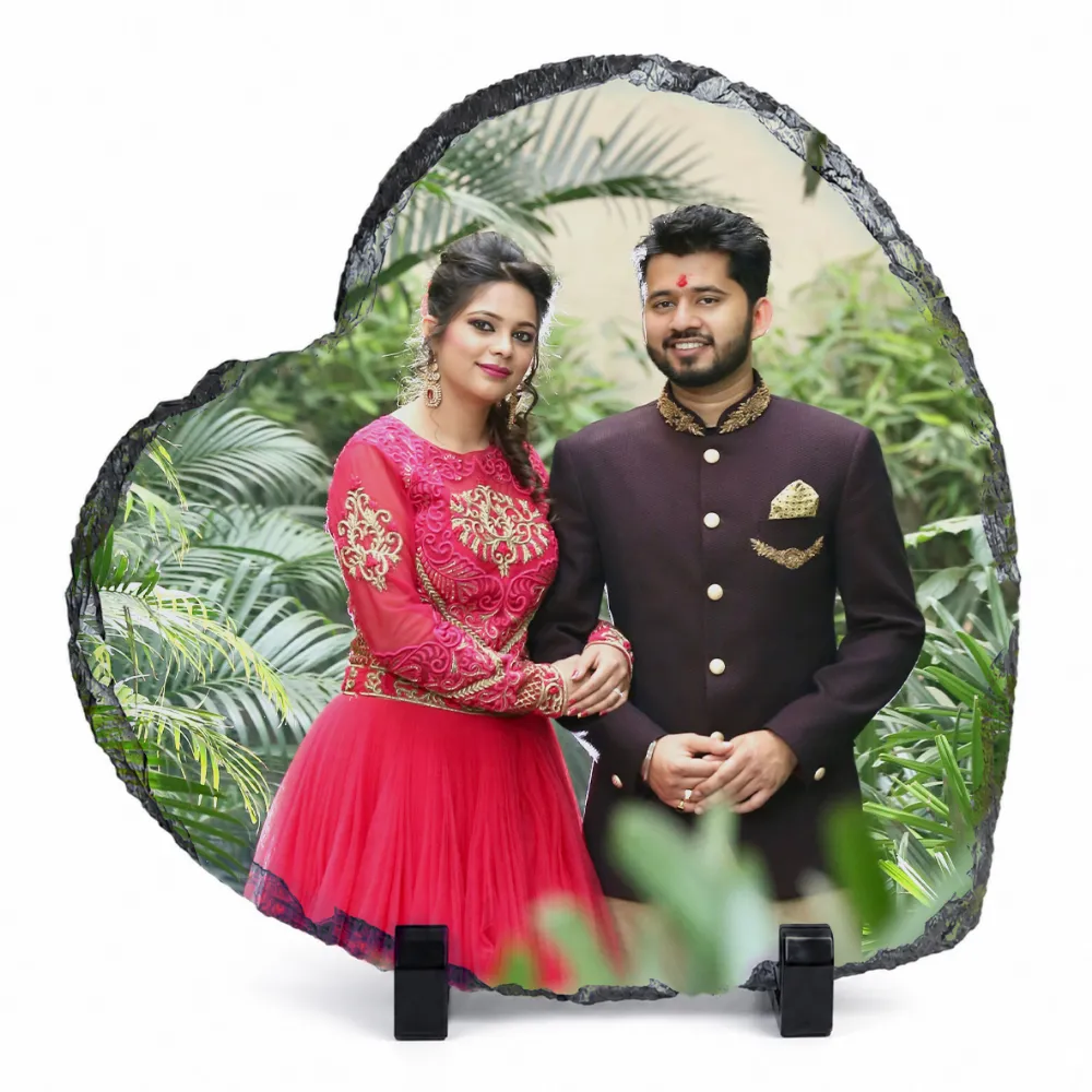 A heart shape natural rock stone frame from upahar18, displaying a personalized custom photo printed on its glossy surface. The printed image features a smiling couple in traditional South Asian wedding attire standing against a lush green garden background, supported by two black stands over a plain white background.