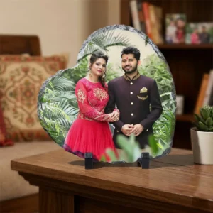 A personalized heart shape rock stone frame from upahar18 placed on a dark wooden table in a living room setup. The custom photo printed on the frame showcases a couple, sitting next to a tiny green plant in a white pot and a decorative black vase.