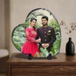 A custom photo printed rock stone frame by upahar18 displayed on a rich wooden drawer cabinet. The heart-shaped personalized frame shows a colorful wedding picture of a couple, sitting next to a black vase with dry flowers against a neutral-toned wall.