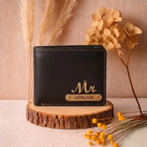 A professional lifestyle product photograph of a personalized black men Wallet from upahar18, placed upright on a rustic wooden log slice placeholder, decorated with dried flowers and grass, highlighting the printed custom name ARNAB and a golden Mr. charm.