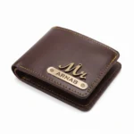An overhead close-up studio shot of a closed personalized deep brown faux Leather Wallet for men from upahar18, featuring a vintage brass-finished 'Mr.' charm and a custom name strip printed with ARNAB, neatly tailored with contrast yellow stitching against a plain white background.