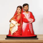 A high-quality printed MDF cutout standee by upahar18 featuring a personalized custom photo of a newlywed Indian couple in traditional red and white Bengali wedding attire, standing back-to-back on a round red base over a wooden surface.