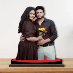 A beautiful printed MDF cutout standee by upahar18 displaying a personalized custom photo of a romantic couple in matching black outfits, with the man holding yellow sunflowers and red roses, standing on a red circular base over a wooden surface.