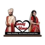 A high-definition printed MDF cutout standee by upahar18 featuring a personalized custom photo of a groom in a cream sherwani and a bride in a red lehenga standing on either side of a heart element printed with the names "Suraj Uma", isolated on a white background.