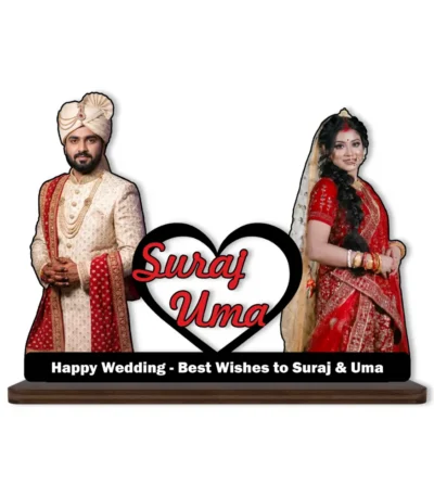 A high-definition printed MDF cutout standee by upahar18 featuring a personalized custom photo of a groom in a cream sherwani and a bride in a red lehenga standing on either side of a heart element printed with the names "Suraj Uma", isolated on a white background.