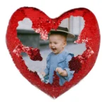 A close-up product shot of a personalized red heart-shaped magic cushion partially revealing a laughing baby's custom photo by upahar18.