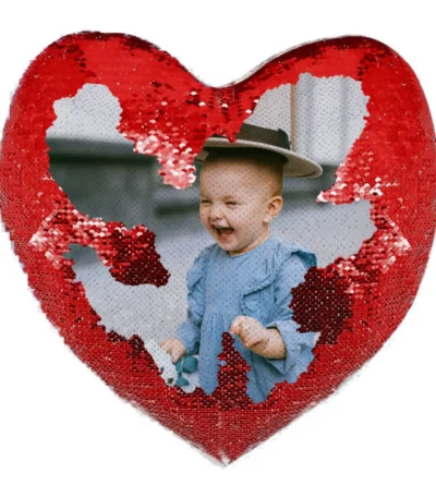 A close-up product shot of a personalized red heart-shaped magic cushion partially revealing a laughing baby's custom photo by upahar18.