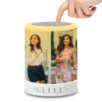A close-up shot of a human hand touching the top silver mesh of the Upahar18 personalized photo printed touch LED Bluetooth speaker. The glowing cylindrical body is fully customized with a vibrant, high-quality photo printed image of two smiling young women outdoors. The smart touch lamp emits a bright yellow ambient glow, beautifully illuminating the customized picture printed on its smooth outer surface.