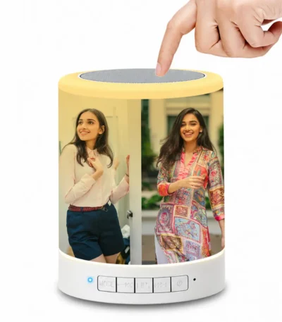 A close-up shot of a human hand touching the top silver mesh of the Upahar18 personalized photo printed touch LED Bluetooth speaker. The glowing cylindrical body is fully customized with a vibrant, high-quality photo printed image of two smiling young women outdoors. The smart touch lamp emits a bright yellow ambient glow, beautifully illuminating the customized picture printed on its smooth outer surface.