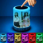 A dark blue studio background highlighting a finger touching the top mesh of the Upahar18 personalized photo printed touch LED Bluetooth speaker, causing it to emit a brilliant cyan blue glow. The customized photo printed image of two women stands out vividly against the light, while a horizontal row of six smaller speakers below showcases the personalized device transitioning through multiple ambient colors.