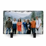 A horizontal rectangle shape natural rock stone frame from upahar18, displaying a personalized custom photo printed on it. The vibrant group photo shows seven friends standing together against a beautiful mountain and pine forest landscape background, supported upright by two black plastic stands on a white background.