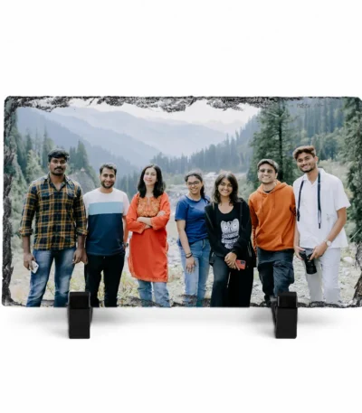 A horizontal rectangle shape natural rock stone frame from upahar18, displaying a personalized custom photo printed on it. The vibrant group photo shows seven friends standing together against a beautiful mountain and pine forest landscape background, supported upright by two black plastic stands on a white background.