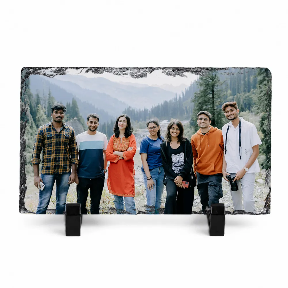 A horizontal rectangle shape natural rock stone frame from upahar18, displaying a personalized custom photo printed on it. The vibrant group photo shows seven friends standing together against a beautiful mountain and pine forest landscape background, supported upright by two black plastic stands on a white background.