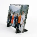 A sharp side angle perspective view of a rectangle shape personalized rock stone frame by upahar18 on a white background. The custom photo printed on the front features a group of friends, while the side angle clearly shows the rugged thickness of the natural rock slate and its sturdy black base stands.
