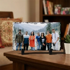 A personalized landscape rectangle rock stone frame from upahar18 styled on a dark wooden table in a living room. The frame features a printed photo of friends in a mountain valley, standing next to a small green plant in a white ceramic pot.