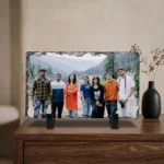 A custom photo printed rock stone frame by upahar18 placed elegantly on a polished wooden cabinet chest. The rectangle personalized stone frame sits next to a black decorative vase with minimal dried flowers under a warm-toned interior setting.