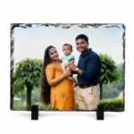 A horizontal rectangle shape natural rock stone frame from upahar18, featuring a personalized custom photo printed on it. The image shows a happy family of three—a mother in a yellow traditional outfit holding a baby, and a father in a dark blue shirt—smiling outdoors with green trees and a cloudy sky in the background, supported by two black stands on a white background.