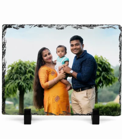 A horizontal rectangle shape natural rock stone frame from upahar18, featuring a personalized custom photo printed on it. The image shows a happy family of three—a mother in a yellow traditional outfit holding a baby, and a father in a dark blue shirt—smiling outdoors with green trees and a cloudy sky in the background, supported by two black stands on a white background.