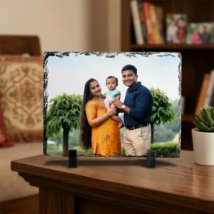A personalized landscape rectangle rock stone frame from upahar18 styled on a dark wooden tabletop in a living room. The frame displays a printed photo of a smiling family, placed neatly beside a minimal green plant in a white ceramic pot.