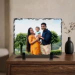 A custom photo printed rock stone frame by upahar18 placed elegantly on a polished wooden chest cabinet. The rectangle personalized frame displays a family portrait, standing next to a decorative black vase with dry flowers under a cozy room setting.