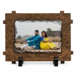 A straight front view of a horizontal rectangle shape natural rock stone frame from upahar18 on a white background. The frame showcases a personalized custom photo printed of a couple sitting back-to-back, highlighted by a dark chiseled stone edge and an outer faux-wooden textured frame border resting on twin black stands.