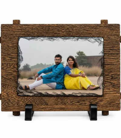A straight front view of a horizontal rectangle shape natural rock stone frame from upahar18 on a white background. The frame showcases a personalized custom photo printed of a couple sitting back-to-back, highlighted by a dark chiseled stone edge and an outer faux-wooden textured frame border resting on twin black stands.
