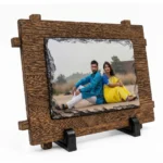 A side angle perspective view of a landscape rectangle shape natural rock stone frame by upahar18 featured within a rustic brown wooden outer frame design. The personalized custom photo printed on the front displays a couple in traditional ethnic attire, sitting outdoors, supported by two black stands against a clean white background.