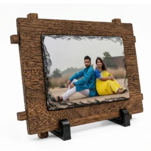 A side angle perspective view of a landscape rectangle shape natural rock stone frame by upahar18 featured within a rustic brown wooden outer frame design. The personalized custom photo printed on the front displays a couple in traditional ethnic attire, sitting outdoors, supported by two black stands against a clean white background.