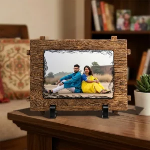 A personalized rectangle rock stone frame from upahar18 displayed on a dark wooden table in a living room backdrop. The frame showcases a custom photo printed of a couple with a textured wooden outer frame border, positioned next to a small green plant in a white pot.