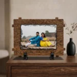 A custom photo printed rock stone frame by upahar18 resting on a polished wooden chest cabinet. The personalized rectangular frame with an outer wood texture border stands next to a decorative black ceramic vase holding dry branches against a neutral wall.