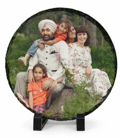 A circular round shape natural rock stone frame from upahar18, showcasing a personalized custom photo printed on it. The image features a smiling family portrait of five members, including a man in a white turban and vest, sitting outdoors on a rock against a green natural background, supported by two black stands on a white background.