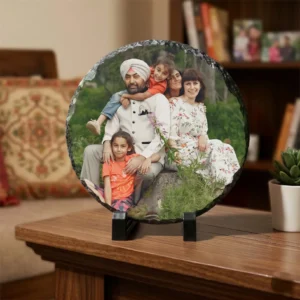 A personalized circular round rock stone frame from upahar18 styled on a polished dark wooden table inside a cozy living room. The frame features a printed photo of a family, placed neatly beside a tiny green succulent in a white pot with a warm blurred background.