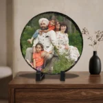 A custom photo printed rock stone frame by upahar18 showcased on a polished wooden drawer cabinet chest. The round personalized frame displays a colorful family photo, standing elegantly next to a decorative black vase with dry flowers under a soft interior setting.