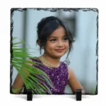 A 1:1 square shape natural rock stone frame from upahar18, showcasing a personalized custom photo printed on it. The vivid print features a sweet smiling young girl in a shimmering purple sequin dress with green palm leaves slightly covering the foreground, supported by two black stands against a clean white background.