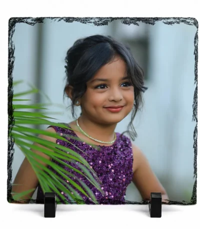 A 1:1 square shape natural rock stone frame from upahar18, showcasing a personalized custom photo printed on it. The vivid print features a sweet smiling young girl in a shimmering purple sequin dress with green palm leaves slightly covering the foreground, supported by two black stands against a clean white background.