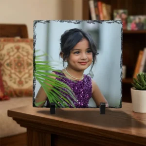 A personalized square rock stone frame from upahar18 styled on a polished dark wooden table in a living room. The frame features a printed photo of a smiling kid, sitting next to a tiny green houseplant in a white pot and a blurred living room interior.