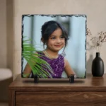 A custom photo printed rock stone frame by upahar18 placed on a polished wooden chest cabinet. The square personalized frame stands elegantly beside a modern black ceramic vase holding dry flowers under a soft home lighting setting.