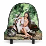 A bell shape natural rock stone frame from upahar18, featuring a personalized custom photo printed on it. The image shows a smiling woman in a light pink dress posing next to a vintage brown and cream scooter in a lush green outdoor setting, placed on two black plastic stands against a clean white background.