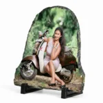 Side angle perspective view of a bell shape personalized rock stone frame by upahar18, showing a custom photo printed on a textured stone slab. The picture features a young woman posing with a classic scooter, highlighting the thickness, natural chiseled borders, and supporting black stands on a white background.