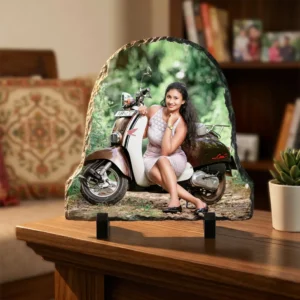 A personalized bell shape rock stone frame from upahar18 displayed on a dark wooden table in a cozy living room. The custom photo printed on the frame shows a woman with a scooter, placed next to a small green plant in a white pot and a decorative black vase.