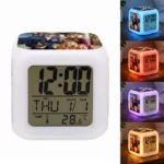 A studio product shot of the Upahar18 personalized photo printed led alarm clock in a square cube shape. The front-facing digital screen clearly displays a black numeric font showing the time as 12:00, day as THU, month and date as 1/1, and temperature as 28.6°C. The top surface features a colorful family photo printed graphic of parents and children in Halloween costumes. On the right side, a vertical stack of four smaller images displays the clock glowing on a wooden table in blue, red, purple, and yellow ambient nightlight modes, illuminating a custom photo printed panel of a family on its side.